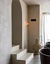 Ljus interiör med valv, små trappsteg, vägglampa och stora dekorativa kärl vid ett fönster.