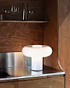 Vit bordslampa på en metallhylla i en träklädd nisch med glas ovanför.