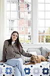 Kvinna sitter på soffa vid stort fönster med en hund bredvid sig i ett ljust rum.