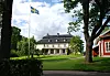 Herrgård med svensk flagga och grön parkmiljö en solig sommardag.
