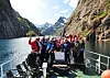 Turistgrupp vinkar på däck under båttur genom smal fjord med höga berg